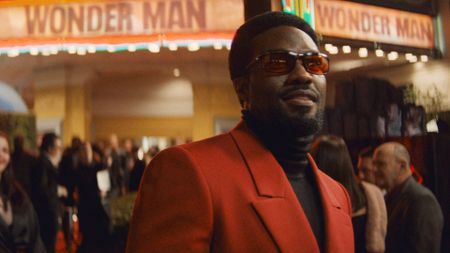 Simon Williams smiling as he stands outside at a movie premiere with sunglasses and a red suit on in Marvel's Wonder Man