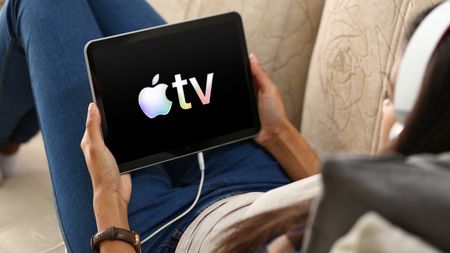 The Apple TV logo on an iPad that someone's holding while sat on a couch